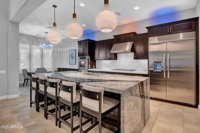 kitchen featuring light stone counters, hanging light fixtures, stainless steel appliances, and a kitchen island with sink