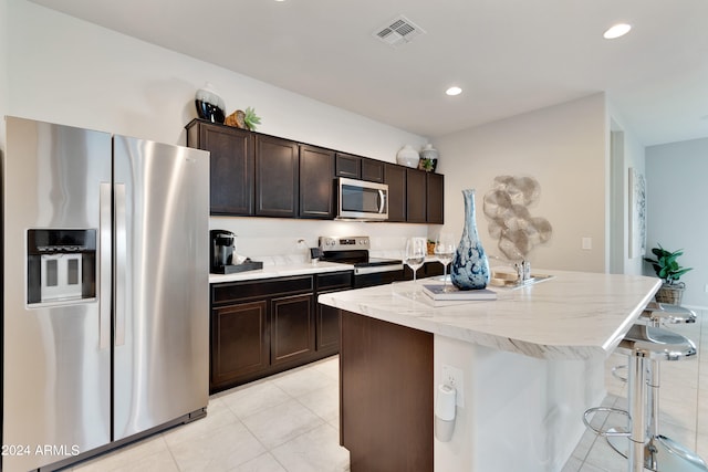 kitchen featuring light tile patterned flooring, appliances with stainless steel finishes, an island with sink, a breakfast bar area, and dark brown cabinets