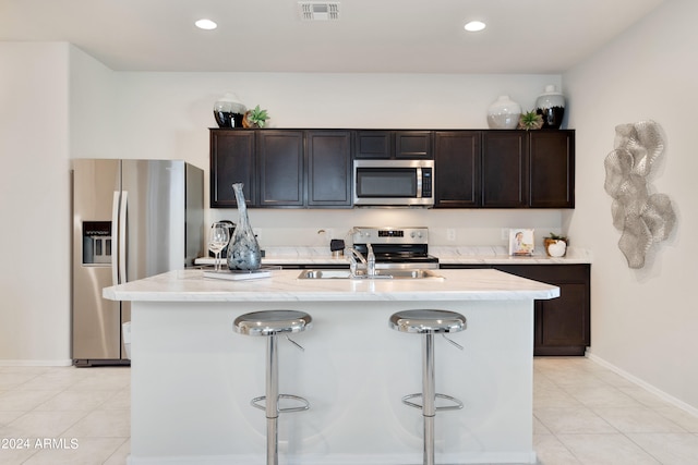 kitchen with light tile patterned flooring, dark brown cabinetry, a center island with sink, appliances with stainless steel finishes, and a kitchen breakfast bar