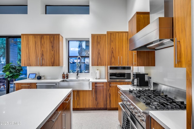 kitchen with light countertops, wall chimney exhaust hood, brown cabinets, a sink, and stainless steel appliances