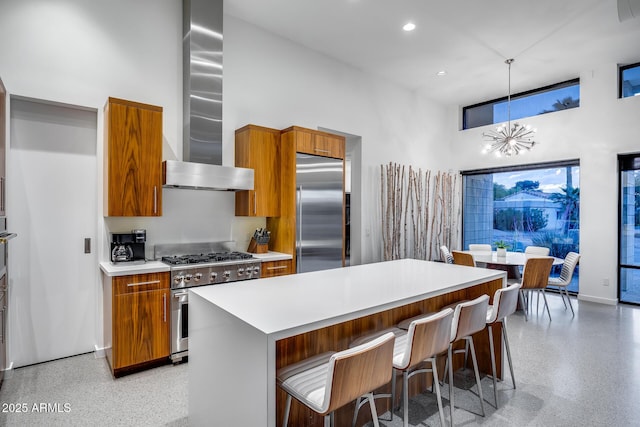 kitchen featuring a towering ceiling, light speckled floor, wall chimney exhaust hood, brown cabinets, and appliances with stainless steel finishes