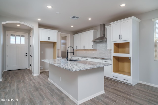 kitchen with a kitchen island with sink, light stone countertops, sink, white cabinets, and wall chimney range hood