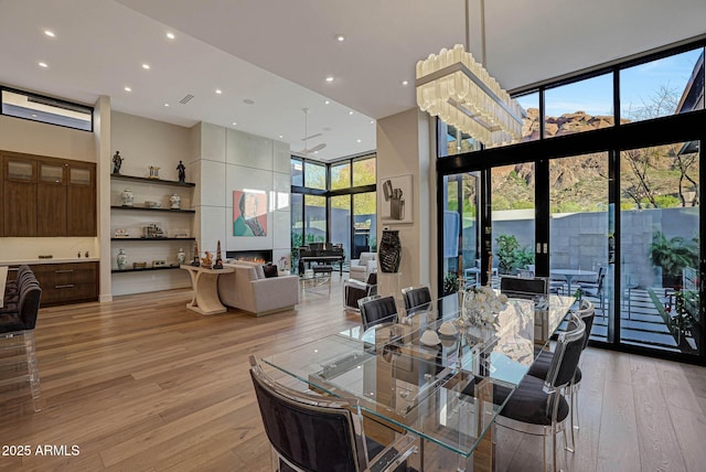 dining room with a large fireplace, light wood finished floors, a high ceiling, a wall of windows, and recessed lighting