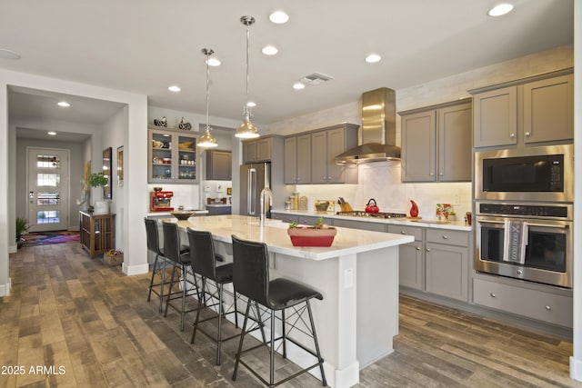 kitchen featuring light countertops, visible vents, appliances with stainless steel finishes, a kitchen island with sink, and wall chimney exhaust hood