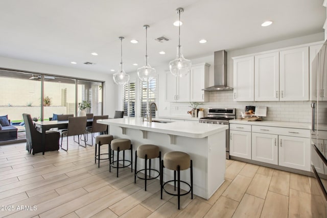 kitchen featuring sink, decorative light fixtures, stainless steel stove, wall chimney exhaust hood, and a center island with sink