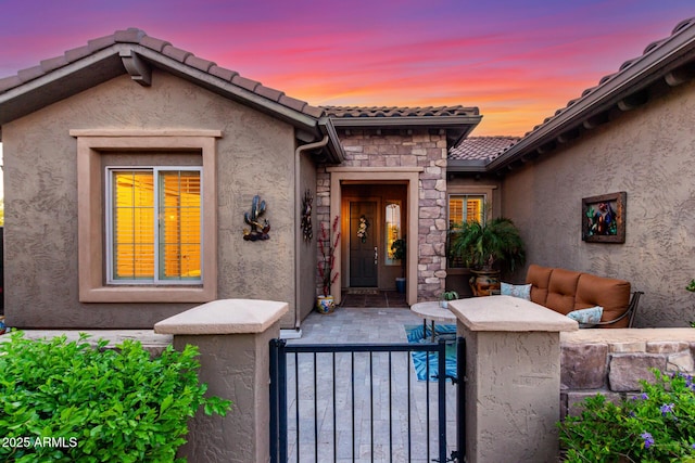exterior entry at dusk with fence, a tile roof, stone siding, stucco siding, and a gate