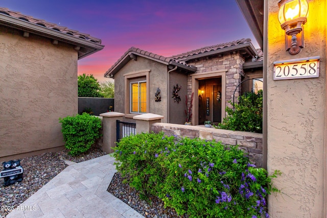 exterior entry at dusk with a tiled roof, stone siding, and stucco siding