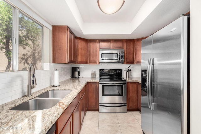 kitchen featuring a tray ceiling, light tile patterned flooring, a sink, appliances with stainless steel finishes, and brown cabinetry