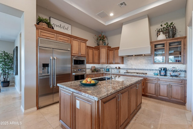 kitchen featuring visible vents, a sink, wall chimney exhaust hood, brown cabinetry, and built in appliances