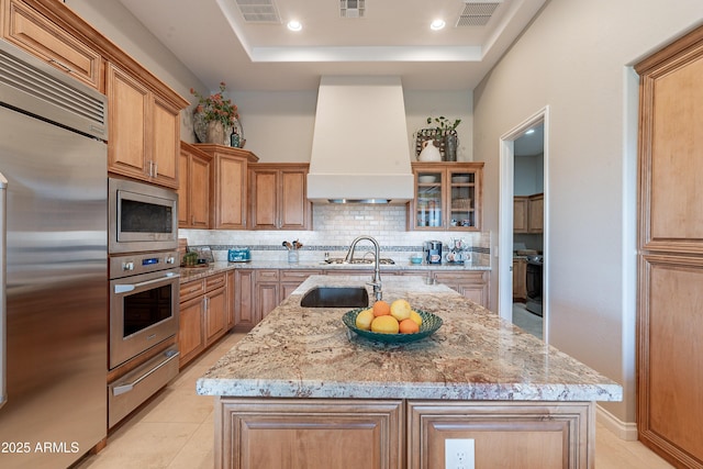 kitchen with a kitchen island with sink, built in appliances, a raised ceiling, a warming drawer, and wall chimney exhaust hood