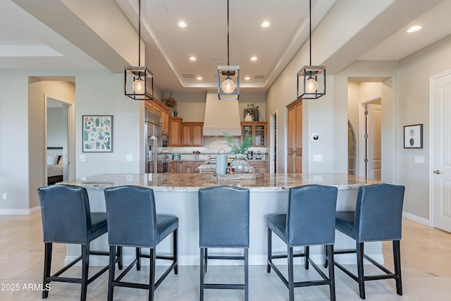 kitchen featuring a kitchen bar, brown cabinets, a tray ceiling, backsplash, and wall chimney range hood