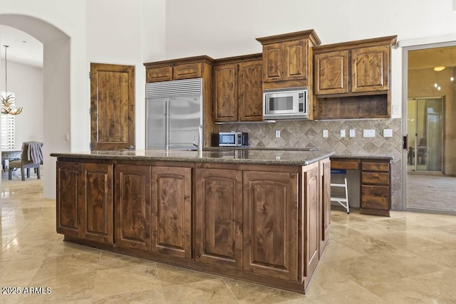 kitchen with a center island with sink, dark stone countertops, tasteful backsplash, arched walkways, and built in appliances