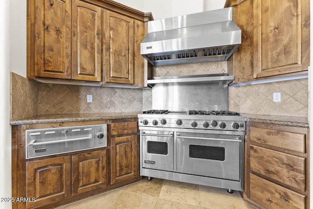 kitchen featuring tasteful backsplash, brown cabinets, wall chimney exhaust hood, and range with two ovens