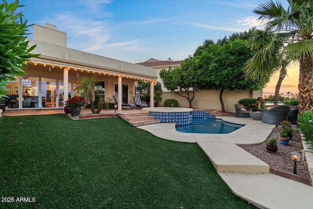 pool at dusk with a lawn, a patio area, fence, a fenced in pool, and a jacuzzi