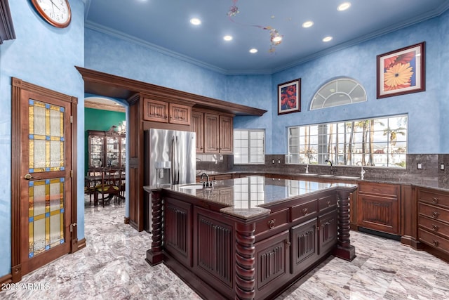 kitchen featuring stainless steel refrigerator with ice dispenser, sink, ornamental molding, dark stone counters, and a kitchen island with sink