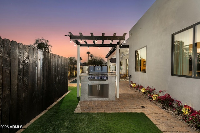 patio terrace at dusk with a yard, grilling area, an outdoor kitchen, a pergola, and a fenced backyard