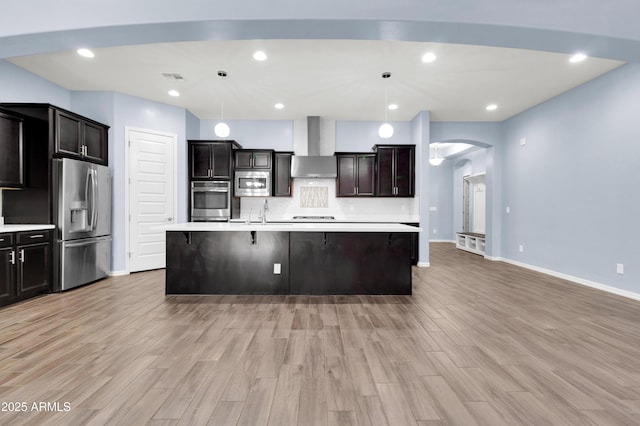 kitchen with stainless steel appliances, wall chimney exhaust hood, light wood finished floors, decorative backsplash, and light countertops