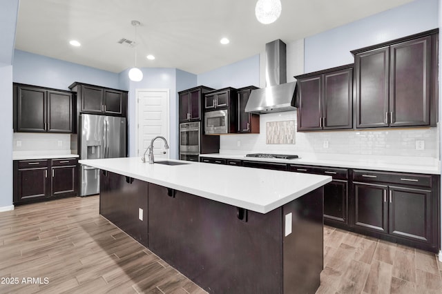 kitchen featuring a center island with sink, stainless steel appliances, decorative light fixtures, wall chimney range hood, and sink