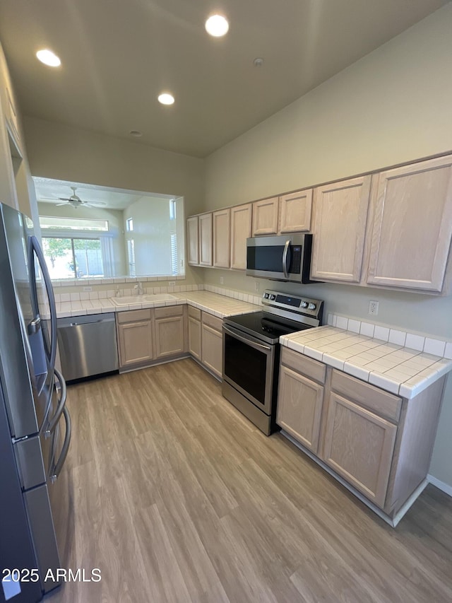 kitchen featuring appliances with stainless steel finishes, tile countertops, light brown cabinetry, sink, and light hardwood / wood-style floors