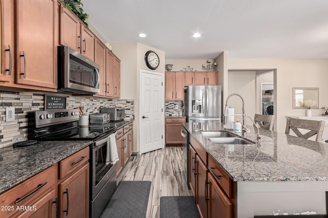 kitchen with a kitchen island with sink, a sink, tasteful backsplash, stainless steel appliances, and brown cabinetry