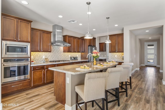 kitchen featuring a kitchen bar, an island with sink, stainless steel appliances, wall chimney exhaust hood, and light stone counters