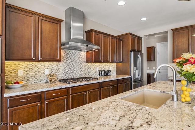 kitchen with light stone countertops, appliances with stainless steel finishes, wall chimney range hood, decorative backsplash, and sink