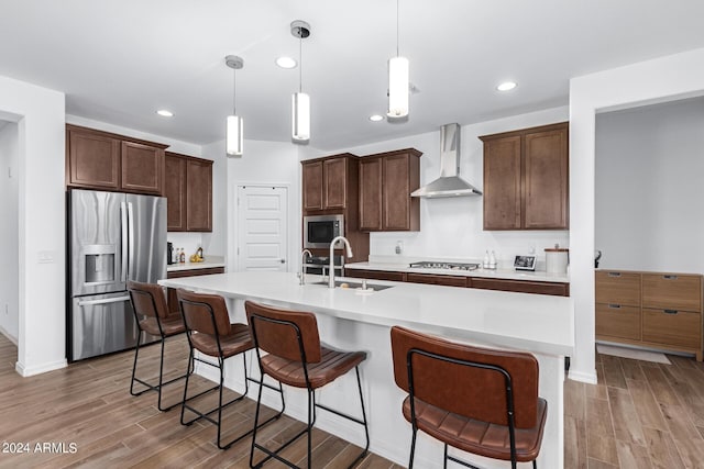 kitchen featuring sink, hanging light fixtures, stainless steel appliances, wall chimney range hood, and a center island with sink