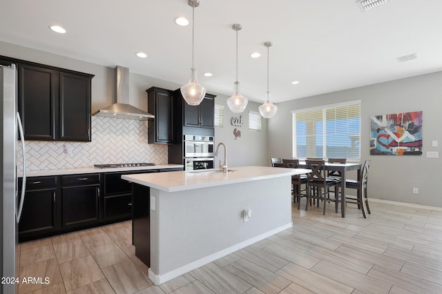 kitchen featuring an island with sink, hanging light fixtures, sink, wall chimney exhaust hood, and stainless steel appliances