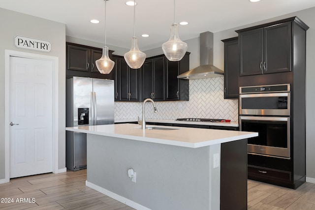 kitchen featuring wall chimney range hood, a kitchen island with sink, pendant lighting, sink, and stainless steel appliances