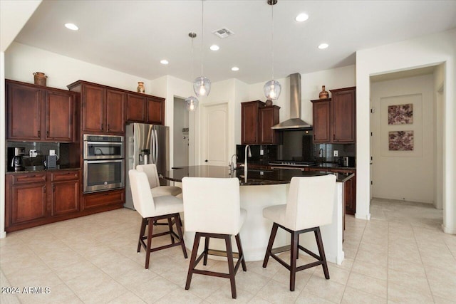 kitchen featuring tasteful backsplash, wall chimney exhaust hood, stainless steel appliances, a kitchen island with sink, and hanging light fixtures