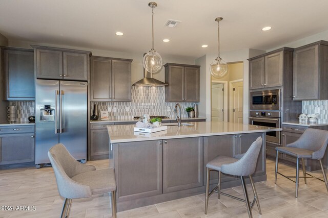 kitchen with stainless steel appliances, light countertops, wall chimney range hood, tasteful backsplash, and a center island with sink