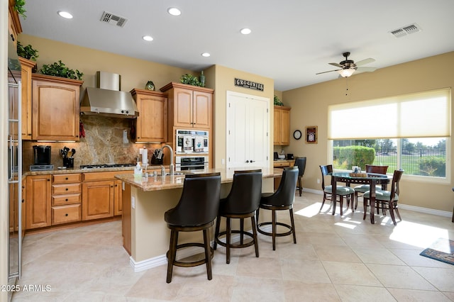 kitchen featuring light stone countertops, gas cooktop, ceiling fan, wall chimney range hood, and an island with sink