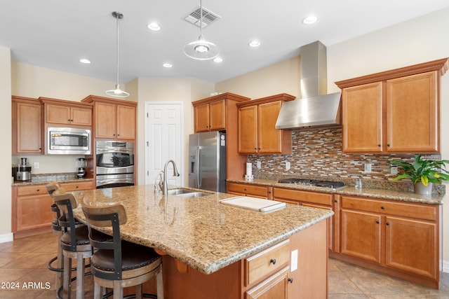 kitchen with stainless steel appliances, sink, wall chimney exhaust hood, an island with sink, and hanging light fixtures