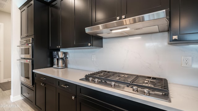 kitchen with decorative backsplash, appliances with stainless steel finishes, dark cabinets, light countertops, and under cabinet range hood