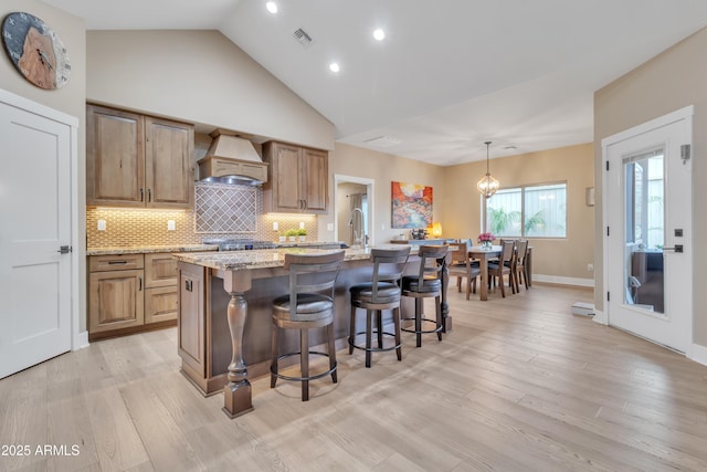 kitchen featuring tasteful backsplash, custom range hood, light wood-style floors, a kitchen island with sink, and a kitchen breakfast bar