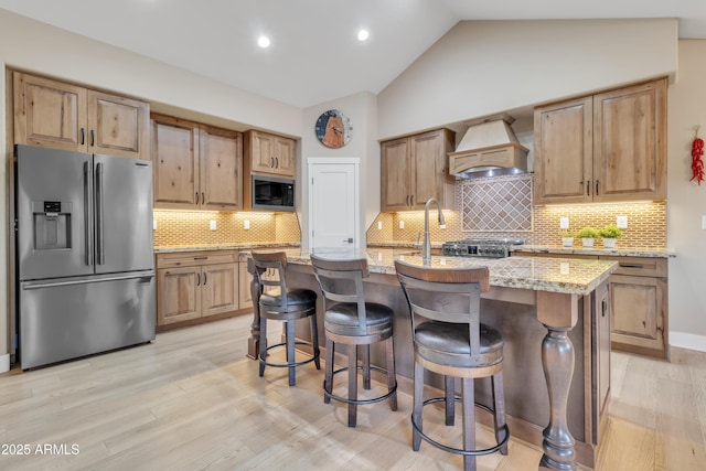 kitchen with stainless steel fridge, a center island with sink, custom range hood, light stone counters, and built in microwave