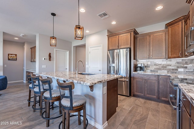 kitchen featuring a kitchen island with sink, pendant lighting, stainless steel appliances, and light stone counters