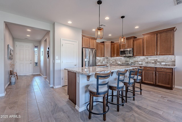 kitchen featuring hanging light fixtures, stainless steel appliances, light stone counters, a kitchen island with sink, and a breakfast bar