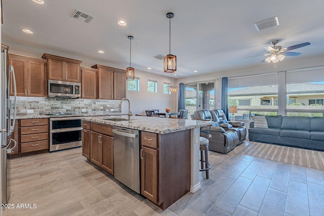 kitchen with light stone counters, stainless steel appliances, sink, hanging light fixtures, and an island with sink