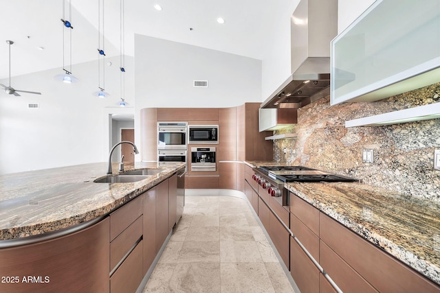 kitchen with sink, wall chimney range hood, stone counters, appliances with stainless steel finishes, and decorative light fixtures