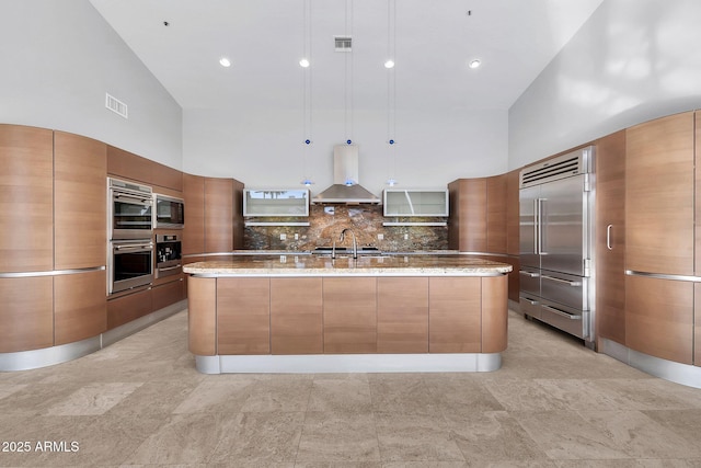 kitchen featuring pendant lighting, built in appliances, a large island with sink, and wall chimney range hood