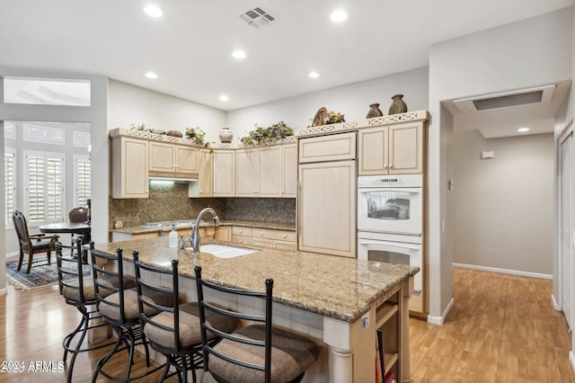 kitchen with light wood-type flooring, light stone counters, a kitchen island with sink, sink, and white double oven