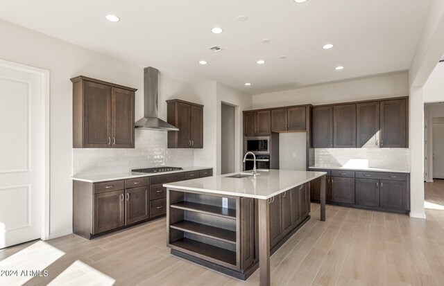 kitchen featuring light hardwood / wood-style floors, wall chimney range hood, appliances with stainless steel finishes, a center island with sink, and backsplash