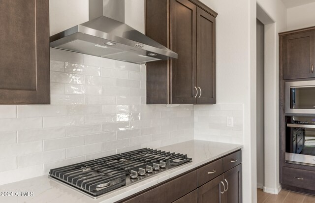 kitchen with light stone countertops, dark brown cabinetry, wall chimney range hood, appliances with stainless steel finishes, and backsplash