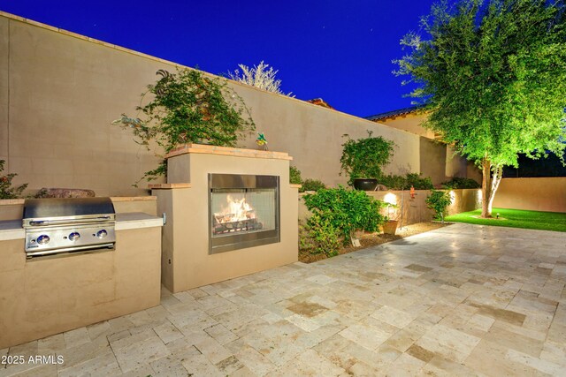 3129 S Honeysuckle Ct, Gold Canyon, AZ 85118 - patio at night featuring fence, exterior kitchen, a lit fireplace, and a grill