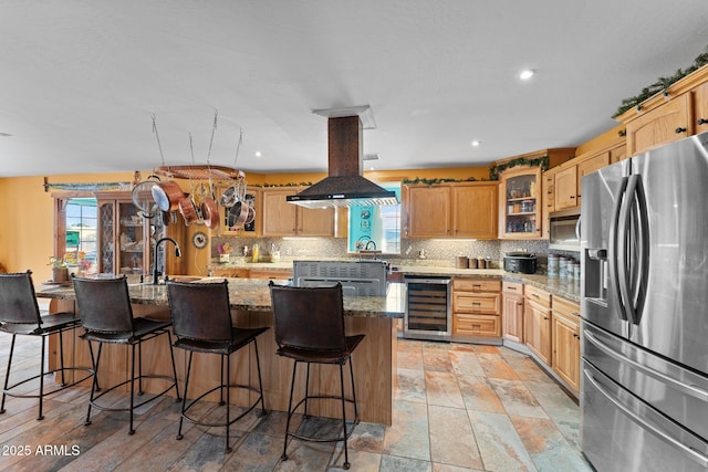 kitchen with island range hood, a center island with sink, stainless steel appliances, beverage cooler, and light stone countertops