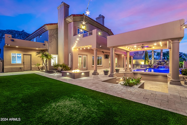 back house at dusk featuring a balcony, ceiling fan, an outdoor fire pit, a patio, and a lawn