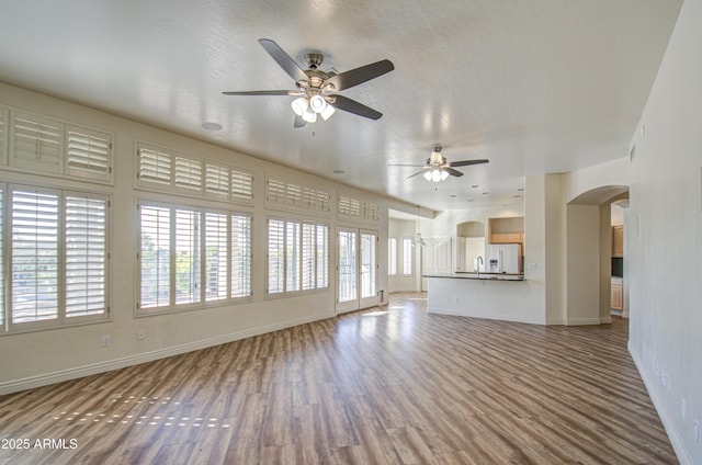 unfurnished living room featuring a ceiling fan, wood finished floors, arched walkways, and baseboards