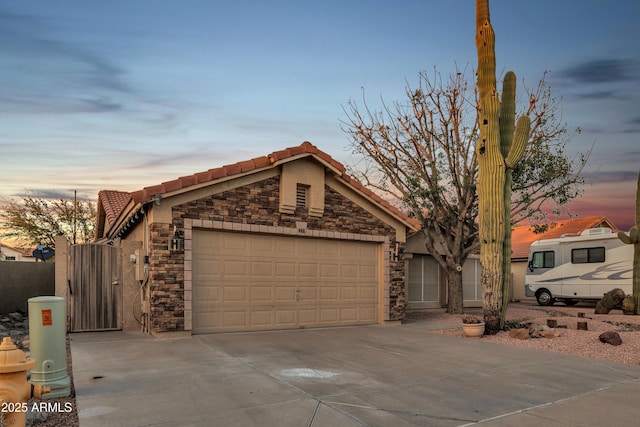 view of front of property featuring a tile roof, a garage, stone siding, driveway, and a gate