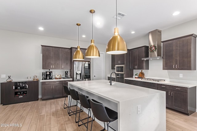 kitchen with a kitchen island with sink, wall chimney range hood, hanging light fixtures, appliances with stainless steel finishes, and light hardwood / wood-style floors